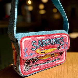May include: A beaded pink and blue handbag with a sardine can design. The bag features the word "SARDINES" in turquoise, along with illustrations of fish and the text "PRODOTTO EN VILLEGGIATURA". The bag has a blue strap.