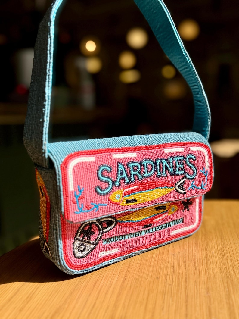 May include: A beaded pink and blue handbag with a sardine can design. The bag features the word "SARDINES" in turquoise, along with illustrations of fish and the text "PRODOTTO EN VILLEGGIATURA". The bag has a blue strap.