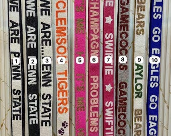 May include: A collection of beaded bag straps in various colors, each displaying a different phrase or team name. The straps include phrases like "We Are Penn State" and "It's Me Hi". The straps are adorned with gold-colored clasps.