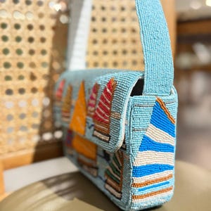 May include: A light blue beaded handbag with a flap closure and a shoulder strap. The bag features colorful beaded sailboat designs in orange, red, and white. The bag is approximately 10 inches wide.