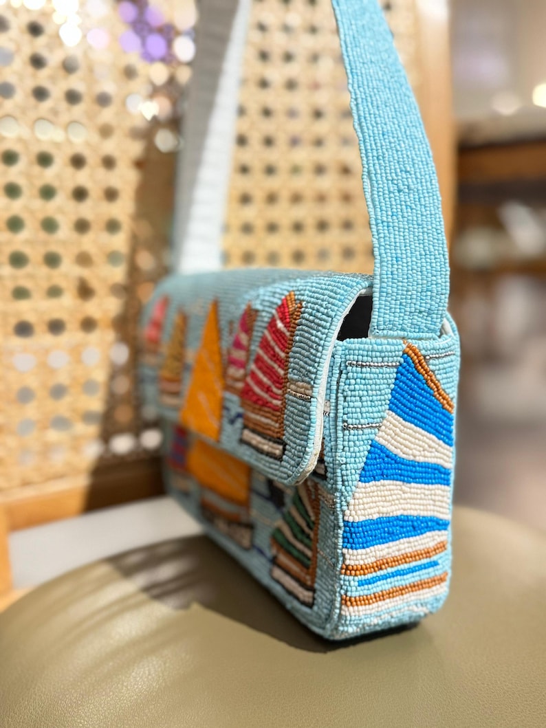 May include: A light blue beaded handbag with a flap closure and a shoulder strap. The bag features colorful beaded sailboat designs in orange, red, and white. The bag is approximately 10 inches wide.