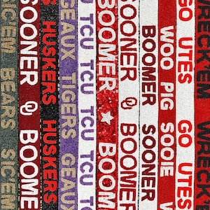 May include: A beaded textile featuring vertical stripes with team slogans and logos. Colors include red, white, black, purple, and green. Phrases like "Boomer Sooner," "Go Utes," and "Sic'em Bears" are visible. The design is likely for sports fans.