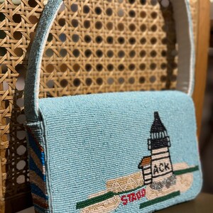 May include: A light blue beaded handbag with a white strap. The bag features a lighthouse design with the text "ACK" and "STAO". The bag has a rectangular shape and a light blue beaded strap.
