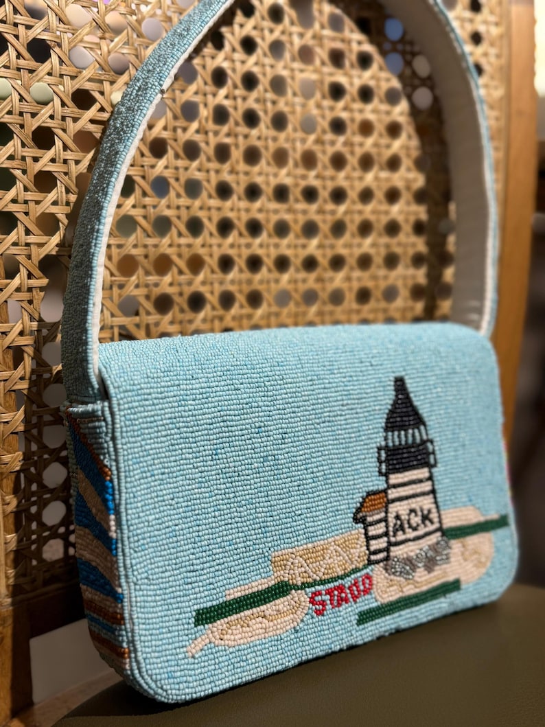 May include: A light blue beaded handbag with a white strap. The bag features a lighthouse design with the text "ACK" and "STAO". The bag has a rectangular shape and a light blue beaded strap.