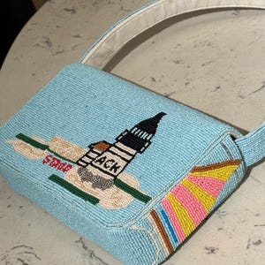 May include: A light blue beaded handbag with a white handle. The bag features a beaded design of a lighthouse with the text "ACK" and "State". The side of the bag has a colorful beaded design.