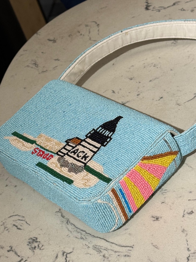 May include: A light blue beaded handbag with a white handle. The bag features a beaded design of a lighthouse with the text "ACK" and "State". The side of the bag has a colorful beaded design.