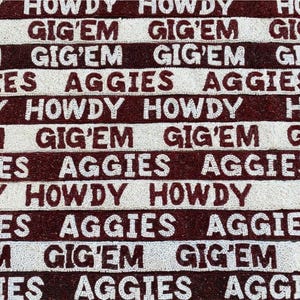 May include: A decorative textile with a repeating pattern of the words "HOWDY", "GIG'EM", and "AGGIES" in white lettering on alternating maroon and white stripes. The design is likely for a college or university.
