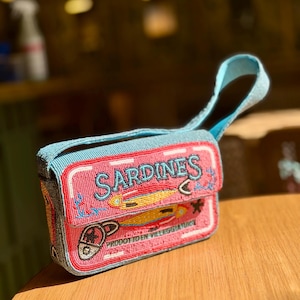 May include: A beaded, rectangular handbag with a blue handle and trim. The bag is pink with the word "SARDINES" in blue, and features illustrations of fish and anchors. The bottom of the bag has the text "PRODOTTO EN VILLEGGIATURA".