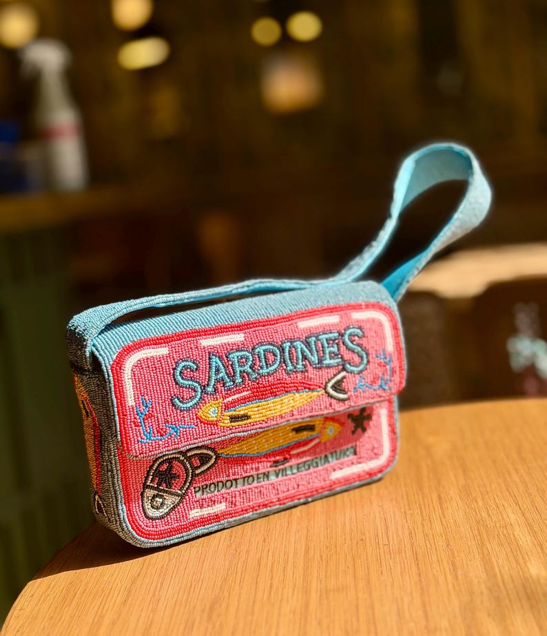 May include: A beaded, rectangular handbag with a blue handle and trim. The bag is pink with the word "SARDINES" in blue, and features illustrations of fish and anchors. The bottom of the bag has the text "PRODOTTO EN VILLEGGIATURA".