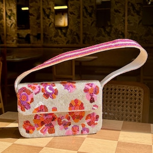 May include: A rectangular handbag with a beaded floral design in shades of pink, orange, and purple. The bag has a white strap and a beaded handle with pink and white stripes. The bag is sitting on a checkered surface.
