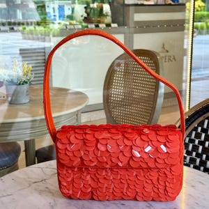 May include: A bright orange rectangular shoulder bag featuring a dense, layered exterior of circular, glossy sequins. The bag has a single, matching orange strap and a structured flap closure that covers the front surface entirely.