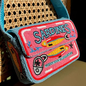 May include: A beaded shoulder bag with a pink front panel featuring the word "SARDINES" in turquoise, along with illustrations of fish. The bag has a turquoise strap and is set against a woven chair.