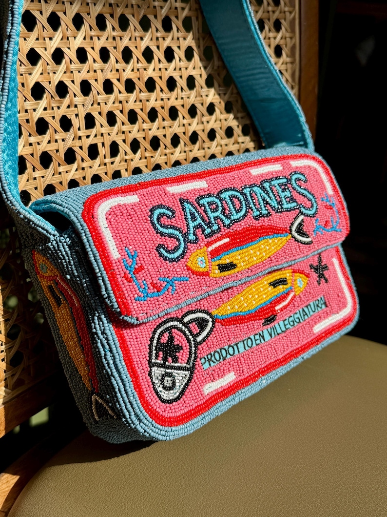 May include: A beaded shoulder bag with a pink front panel featuring the word "SARDINES" in turquoise, along with illustrations of fish. The bag has a turquoise strap and is set against a woven chair.