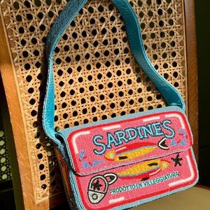 Op de afbeelding: Een geborduurde handtas met een roze achtergrond en een blauwe band. De tas heeft het woord "SARDINES" in blauw, samen met illustraties van vissen en de tekst "PRODOTTO EN VILLEGGIATURA". De tas ligt op een houten stoel.