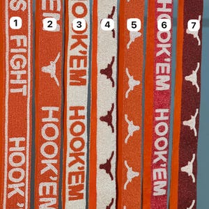 Może przedstawiać: Kolekcja koralikowych wstążek w odcieniach pomarańczu, bieli i brązu. Wstążki zawierają napisy "HOOK 'EM" i "KAS FIGHT", a także sylwetkę longhornu. Wstążki są prawdopodobnie używane do rękodzieła lub jako akcesoria.