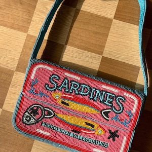 May include: A beaded rectangular handbag with a pink background and the word "SARDINES" in white. The bag features a design of two yellow fish and the text "PRODOTTO EN VILLEGGIATURA". The strap is blue.