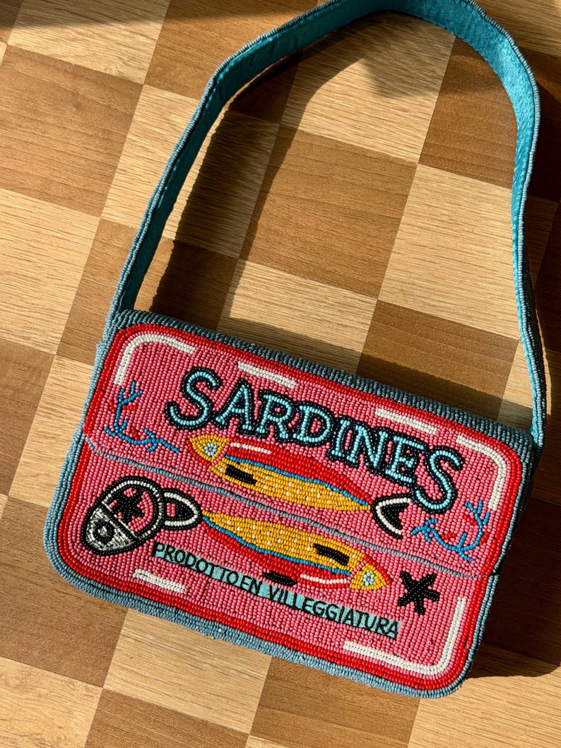 May include: A beaded rectangular handbag with a pink background and the word "SARDINES" in white. The bag features a design of two yellow fish and the text "PRODOTTO EN VILLEGGIATURA". The strap is blue.