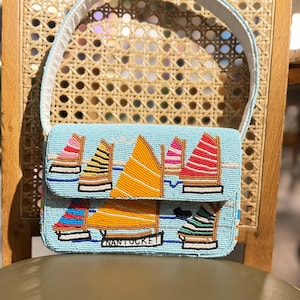 May include: A beaded light blue handbag with a white handle. The bag features a design of colorful sailboats and the word "NANTUCKET" in white. The sailboats have orange, pink, and striped sails. The bag is on a wooden chair.