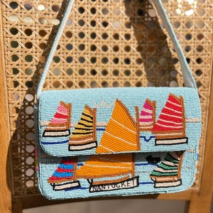 May include: A light blue beaded handbag with a sailboat design. The bag features colorful sailboats with orange, pink, yellow, and red sails. The word "Nantucket" is displayed on the front. The bag has a light blue strap.