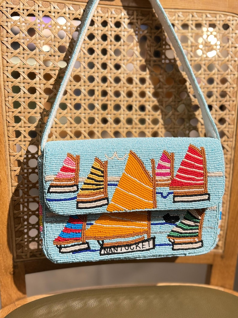 May include: A light blue beaded handbag with a sailboat design. The bag features colorful sailboats with orange, pink, yellow, and red sails. The word "Nantucket" is displayed on the front. The bag has a light blue strap.