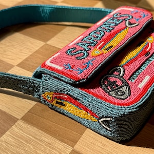 May include: A beaded pink purse with the word "Sardines" in red lettering. The purse features a blue strap and is decorated with yellow and red fish designs. The purse is on a wooden surface.