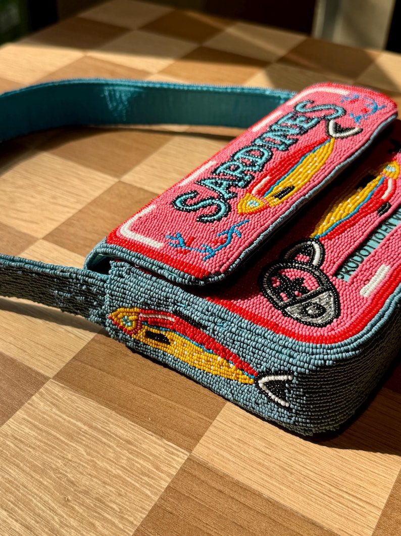 May include: A beaded pink purse with the word "Sardines" in red lettering. The purse features a blue strap and is decorated with yellow and red fish designs. The purse is on a wooden surface.
