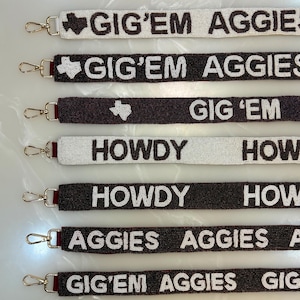 Könnte beinhalten: Eine Auswahl an bestickten Taschenriemen in verschiedenen Farben, mit den Schriftzügen "GIG'EM AGGIES" und "HOWDY" in Weiß. Jeder Riemen hat goldfarbene Verschlüsse und ein Texas-Design.