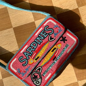 May include: A rectangular beaded purse with a pink background and the word "SARDINES" in blue. The purse features a design of yellow fish and the text "PRODOTTO EN VILLEGGIATURA". The strap is light blue.