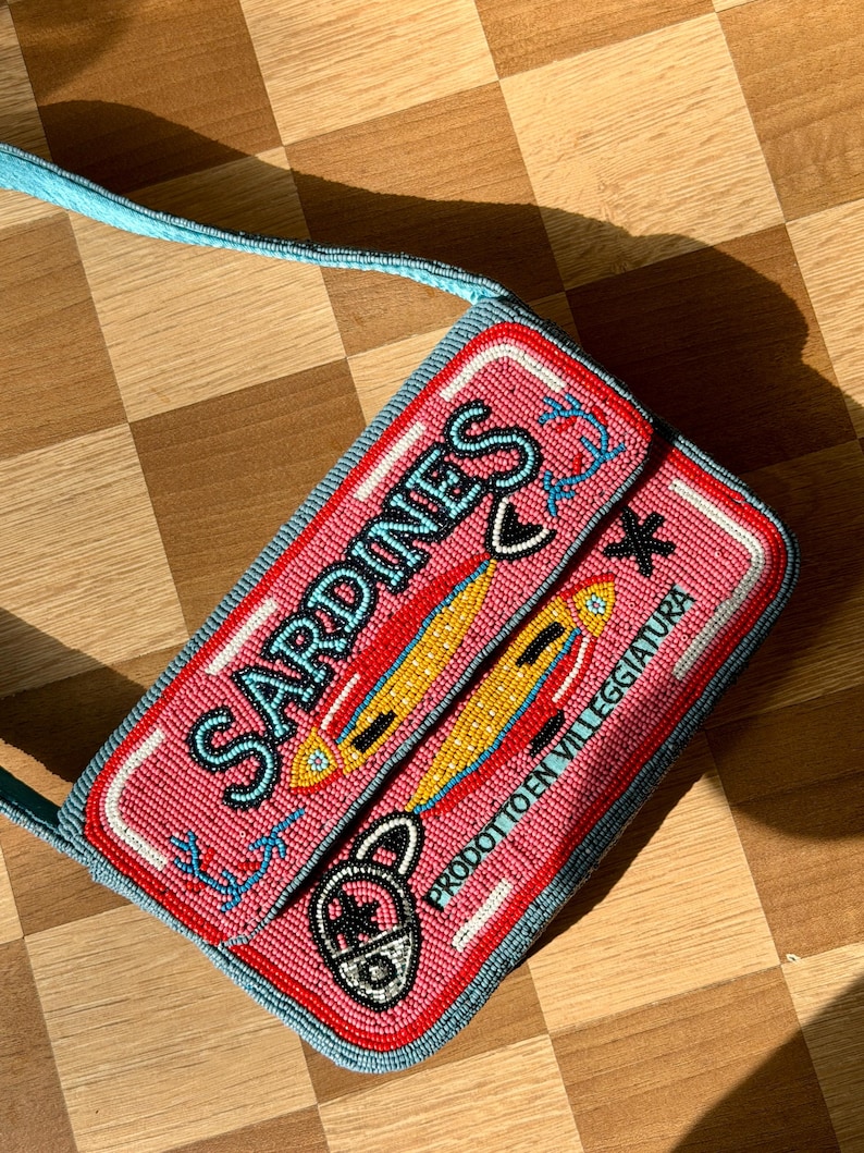 May include: A rectangular beaded purse with a pink background and the word "SARDINES" in blue. The purse features a design of yellow fish and the text "PRODOTTO EN VILLEGGIATURA". The strap is light blue.