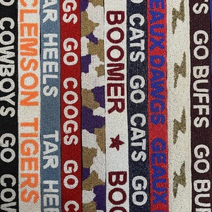 Puede incluir: Un textil bordado con rayas verticales que muestran nombres de equipos y frases. Los colores incluyen negro, naranja, azul, rojo y blanco. El texto incluye "GO COWBOYS", "CLEMSON TIGERS", "TAR HEELS", "COOGS GO COOGS", "BOOMER", "GO CATS", "GEAUX DAWGS" y "GO BUFFS".