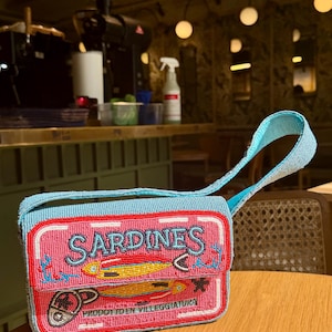 May include: A rectangular, beaded handbag with a light blue strap and trim. The bag features a pink front panel with the word "SARDINES" and illustrations of fish. The text "PRODOTTO EN VILLEGGIATURA" is also visible.