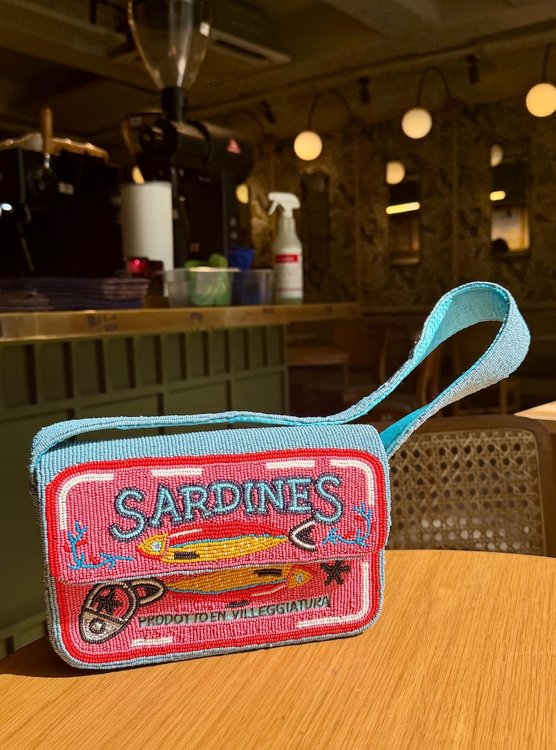 May include: A rectangular, beaded handbag with a light blue strap and trim. The bag features a pink front panel with the word "SARDINES" and illustrations of fish. The text "PRODOTTO EN VILLEGGIATURA" is also visible.