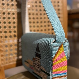 May include: A light blue beaded shoulder bag with a cityscape design. The bag features a long strap and a flap closure. The side of the bag has a colorful sailboat design with pink, yellow, and brown beads.