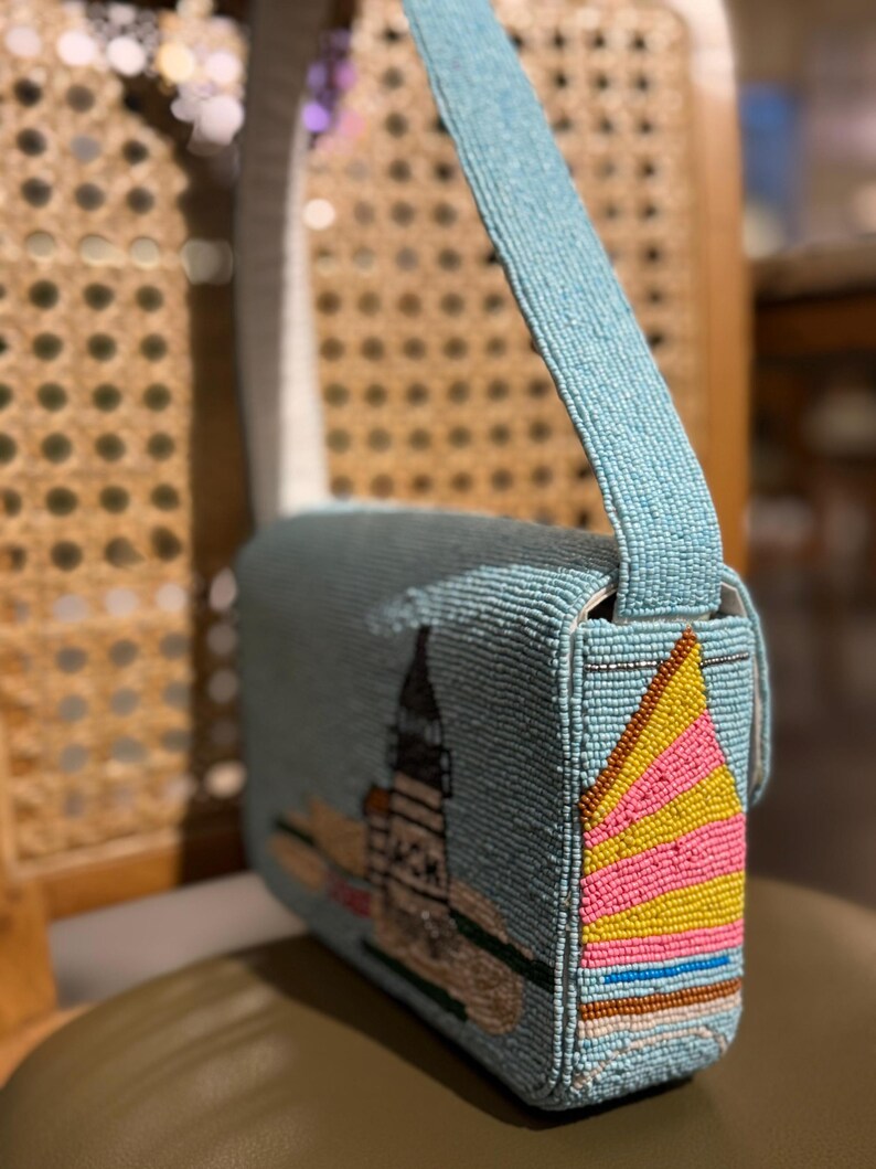 May include: A light blue beaded shoulder bag with a cityscape design. The bag features a long strap and a flap closure. The side of the bag has a colorful sailboat design with pink, yellow, and brown beads.
