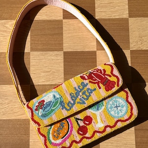 Op de afbeelding: Een gele, met kralen versierde handtas met een roze band. De tas heeft een gestreept patroon en is versierd met kleurrijke kralenafbeeldingen van een kreeft, kersen en andere ontwerpen. De woorden "Dolce Vita" staan op de flap.