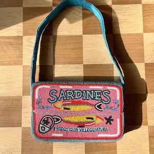 May include: A beaded handbag resembling a sardine can. The pink bag features the word "SARDINES" in turquoise, with illustrations of fish and the text "PRODOTTO EN VILLEGGIATURA". The strap and trim are turquoise.