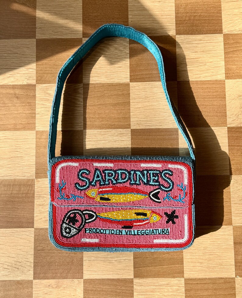 May include: A beaded handbag resembling a sardine can. The pink bag features the word "SARDINES" in turquoise, with illustrations of fish and the text "PRODOTTO EN VILLEGGIATURA". The strap and trim are turquoise.