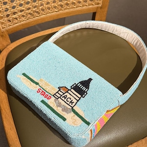 May include: A light blue beaded handbag with a white handle. The bag features a lighthouse design with the word "ACK" and "STUDS" on the front. The bottom of the bag has a colorful striped pattern.