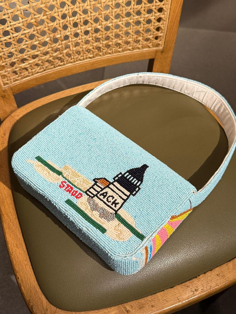 May include: A light blue beaded handbag with a white handle. The bag features a lighthouse design with the word "ACK" and "STUDS" on the front. The bottom of the bag has a colorful striped pattern.