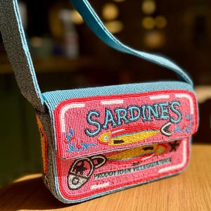 May include: A beaded crossbody bag with a pink sardine can design. The bag features the word "SARDINES" in blue beads, along with illustrations of fish and a can opener. The strap and top of the bag are light blue.
