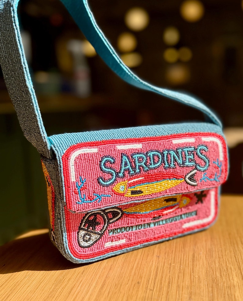 May include: A beaded crossbody bag with a pink sardine can design. The bag features the word "SARDINES" in blue beads, along with illustrations of fish and a can opener. The strap and top of the bag are light blue.