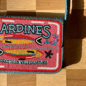 May include: Pink beaded crossbody bag with the word "SARDINES" in turquoise. The bag features two fish illustrations and the text "PRODOTTO EN VILLEGGIATURA" in turquoise. The bag has a red border and a blue strap.