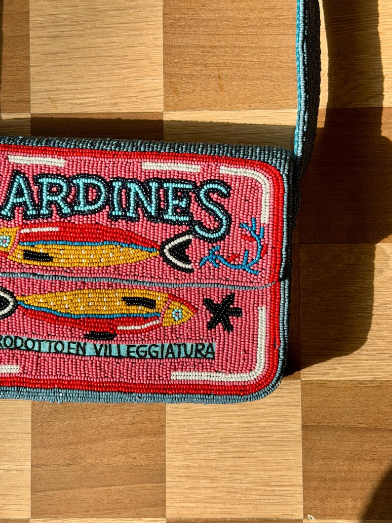 May include: Pink beaded crossbody bag with the word "SARDINES" in turquoise. The bag features two fish illustrations and the text "PRODOTTO EN VILLEGGIATURA" in turquoise. The bag has a red border and a blue strap.