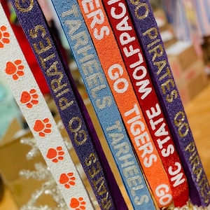 Könnte beinhalten: Eine Sammlung von Perlen-Lanyards in verschiedenen Farben, darunter Weiß, Lila, Blau, Orange und Rot. Die Lanyards zeigen Texte wie "GO PIRATES", "TARHEELS" und "WOLFPACK". Einige haben Pfotenabdruck-Designs.