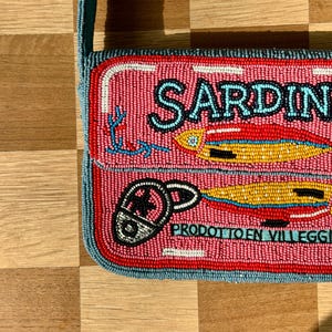 May include: Pink beaded crossbody bag with a sardine can design. The bag features the word "SARDINE" in black letters, along with illustrations of sardines and a can opener. The bag has a teal strap and trim.