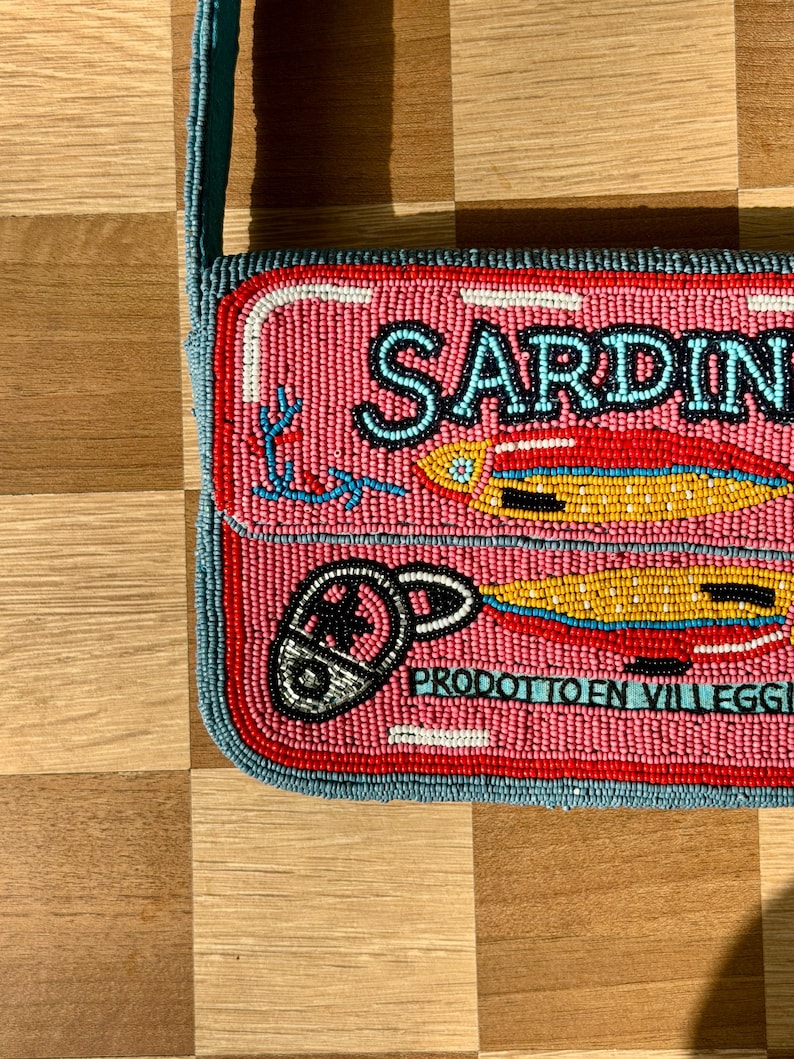 May include: Pink beaded crossbody bag with a sardine can design. The bag features the word "SARDINE" in black letters, along with illustrations of sardines and a can opener. The bag has a teal strap and trim.