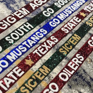 May include: Close-up of several beaded ribbons with team names and phrases like "Gig'Em Aggies", "Go Mustangs", and "Go Oilers". The ribbons are in various colors, including maroon, green, and blue, with white and gold lettering.