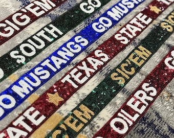 May include: A selection of beaded ribbons, each displaying team names and slogans such as "Gig'Em Aggies" and "Go Mustangs". The ribbons are in shades of maroon, green, and blue, with white and gold lettering. The background is a patterned fabric.