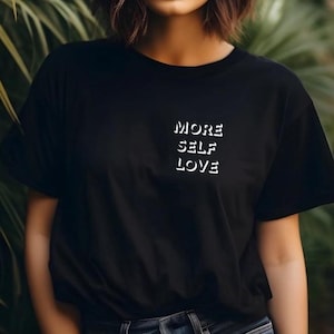 Könnte beinhalten: Schwarzes T-Shirt mit den Worten "MORE SELF LOVE" in Weiß aufgedruckt. Der Text ist vertikal auf der linken Brustseite des T-Shirts angeordnet. Das T-Shirt hat eine einfarbige schwarze Farbe.