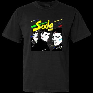 Op de afbeelding: Zwart t-shirt met een grafisch ontwerp. Het ontwerp toont de woorden "Soda Stereo" in het geel, met kleurrijke lijnen. Het bevat ook drie zwart-wit portretten van mensen.